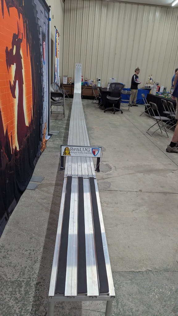 A long gravity-powered LEGO® racing track set up for the RenLUG Royal Rally Racing event, featuring a smooth surface with black strips and a backdrop of a colorful mural.