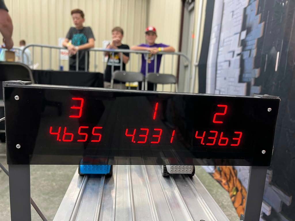 Digital scoreboard displaying race results with times for three LEGO cars, with spectators in the background.