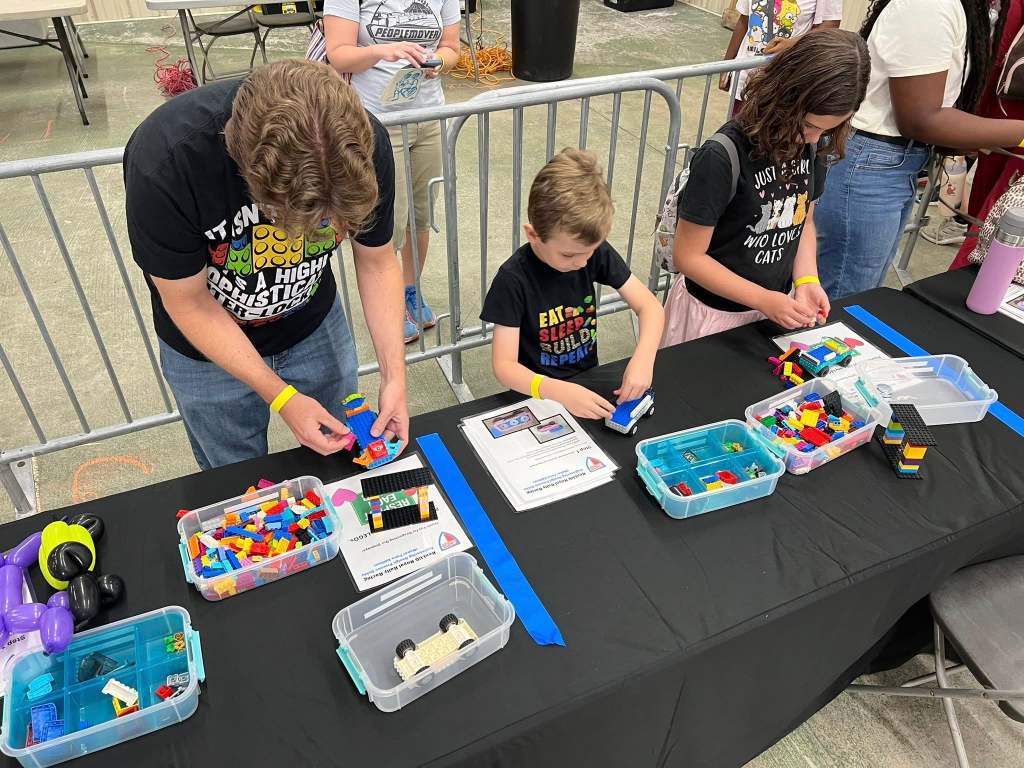 Children and adults participate in a hands-on LEGO® building activity, creating cars from colorful LEGO® bricks at a community event.