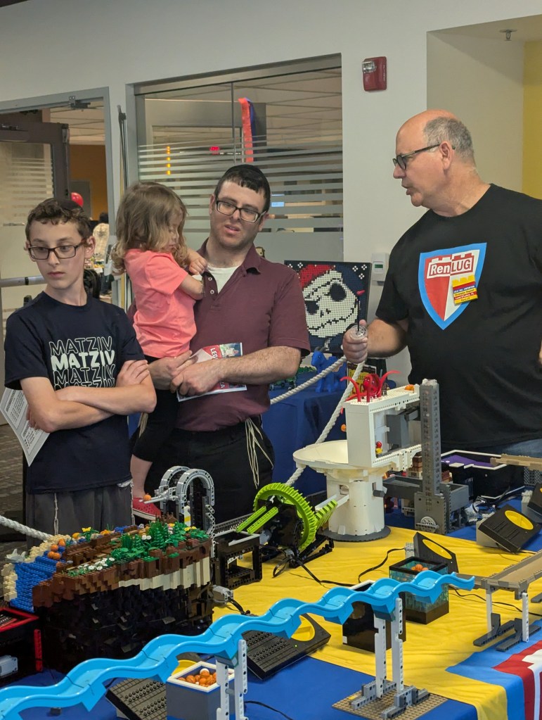 A group of three people, including a young boy and a toddler, are observing a LEGO display at an event. One adult is holding the toddler, while another adult, wearing a 'RenLUG' shirt, is speaking. The display features various LEGO creations and is set up on a table covered with a blue tablecloth.