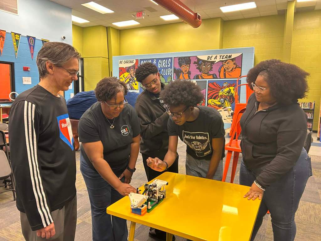 A diverse group of five individuals, including both adults and teenagers, gathered around a yellow table engaged in a robotics project, demonstrating collaboration and learning in a colorful classroom setting.