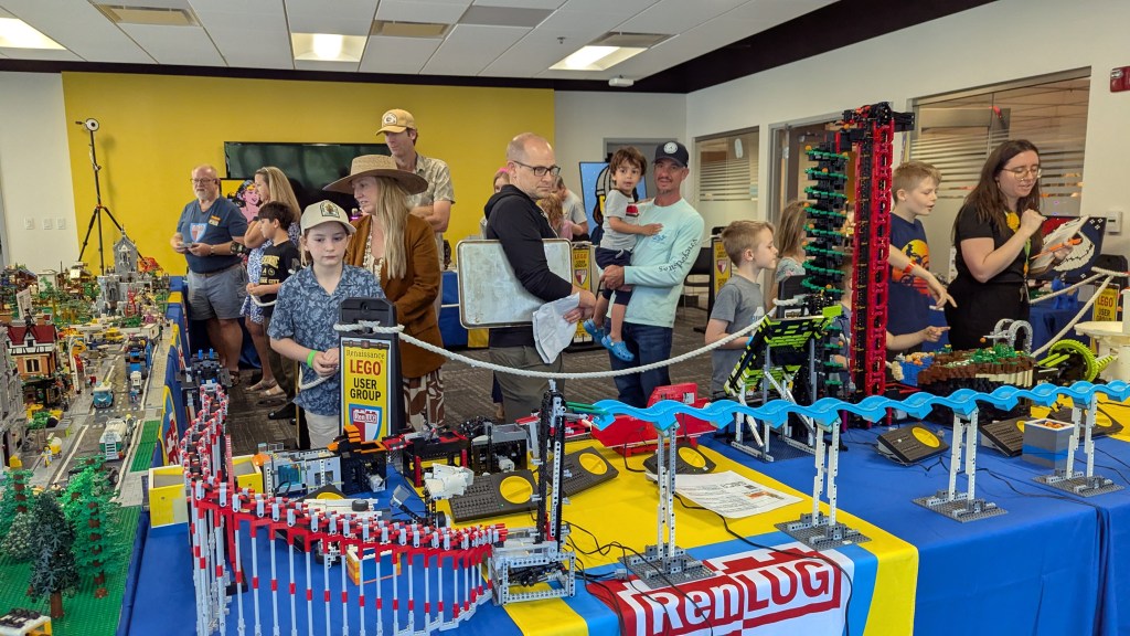 A group of people, including children and adults, viewing a colorful display of LEGO creations at an event. The scene features various LEGO models, including a roller coaster and miniature buildings, set on tables in a large room.